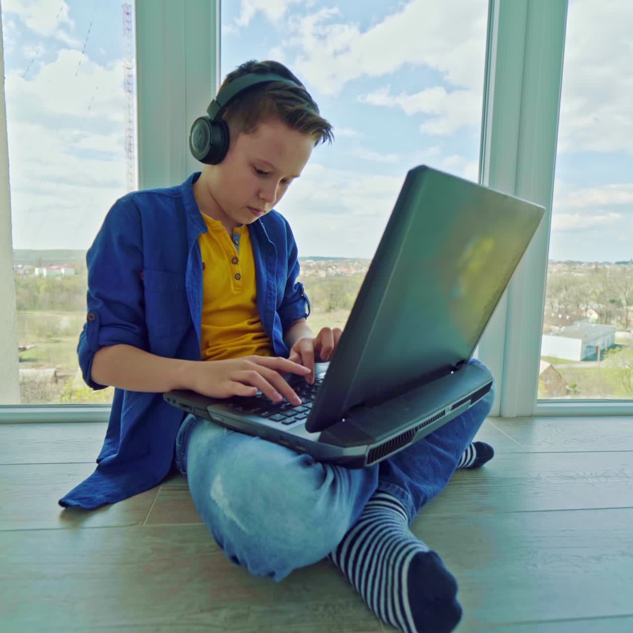 Boy in headphones playing video game on laptop near the window. Teenager gravely looking to the notebook screen pressing the keyboard. Technology concept.