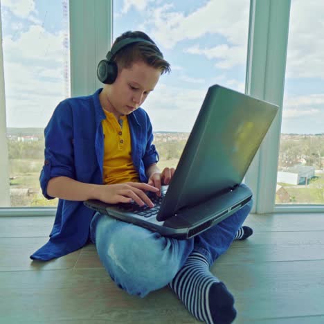 boy in headphones playing video game on laptop near the window. teenager gravely looking to the notebook screen pressing the keyboard. technology concept.