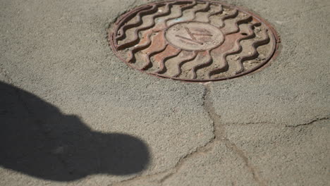 close view of a manhole cover on a cracked asphalt road, partially obscured by a shadow, with people passing by and there shadows casted on the ground