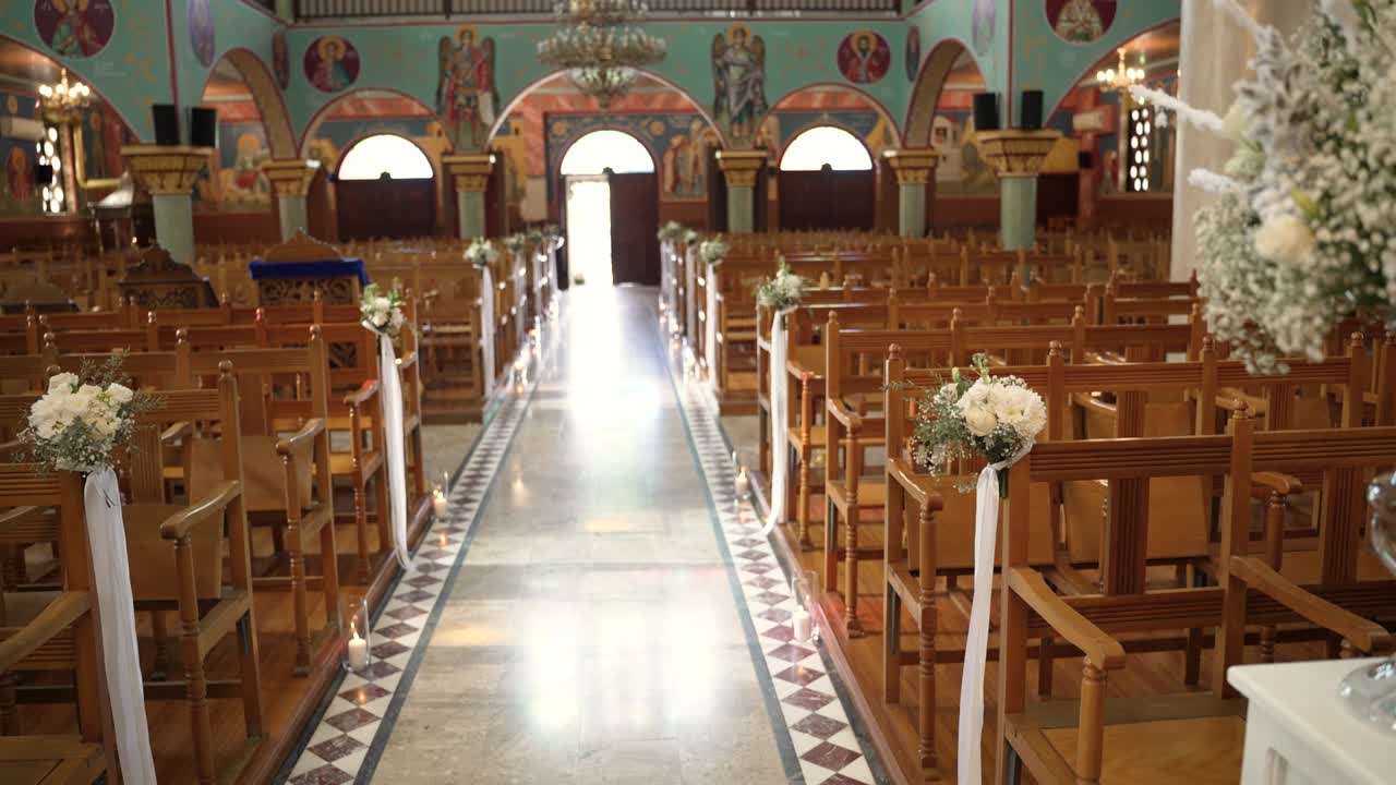 Una cerimonia di matrimonio si svolge in una chiesa. La chiesa ha molte sedie di legno disposte in file. Decorazioni floreali sono legate alle sedie. La luce entra dalle porte principali.