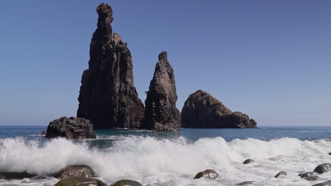 A wave crashes over a rocky beach, with rocks rising from the water in the background. The clear blue sky creates a peaceful yet powerful coastal scene filled with natural beauty.