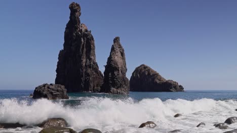 a wave crashes over a rocky beach, with rocks rising from the water in the background. the clear blue sky creates a peaceful yet powerful coastal scene filled with natural beauty.