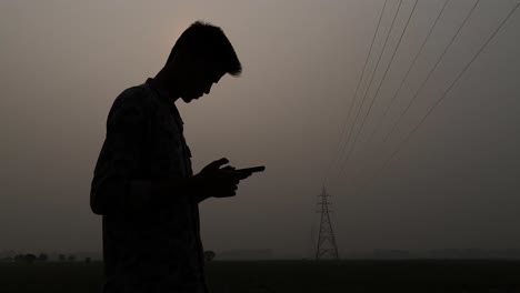 Silueta-De-Un-Joven-Indio-Haciendo-Una-Llamada-En-Un-Teléfono-Inteligente-Con-Un-Pilón-De-La-Línea-Eléctrica-Del-Amanecer-Brumoso-En-Un-Horizonte-Distante