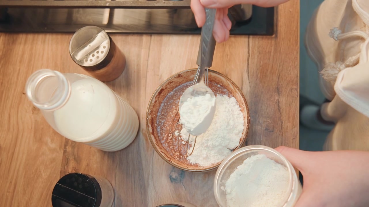 Tilsetning av mel til sjokoladerøre, oversiktsbilde viser melkeflaske, visp og krukke på trebenk, hender blander tørre ingredienser inn i kakaobase, lyst naturlig lys, matlagingsdemonstrasjon i veiledningsstil