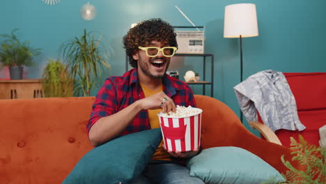 indian man sits on sofa eating popcorn and watching interesting tv serial sport game online at home