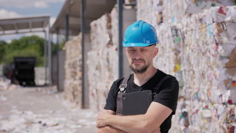 Man-in-hard-hat-and-overalls-staring-at-camera,-paper-recycling-plant