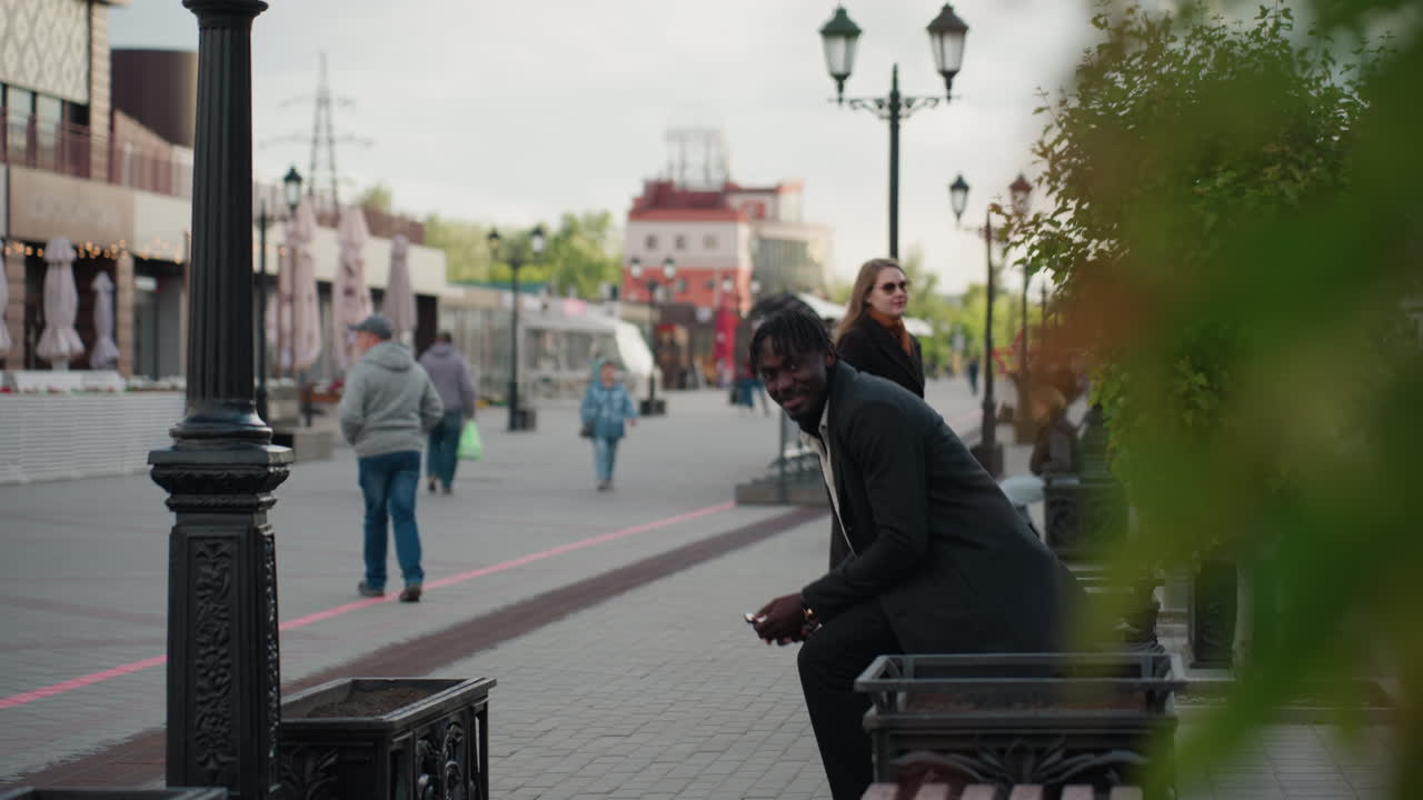 American lady returns phone to her partner and walks past with smile on bright street, surrounded by benches, street lamps, and greenery, creating scene filled with warmth, emotion, and connection