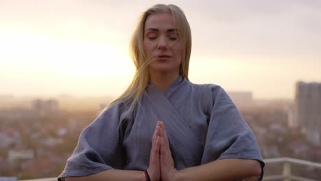 peaceful blonde woman standing on terrace, holding her hands in a namaste gesture