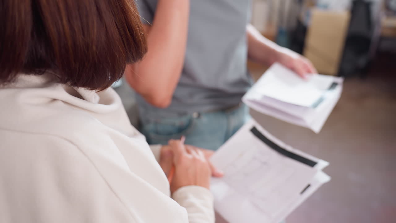 closeup view of two dress makers actively working together during cloth pattern creation session, one sketching design on paper while other describes adjustments using hand gestures during teamwork
