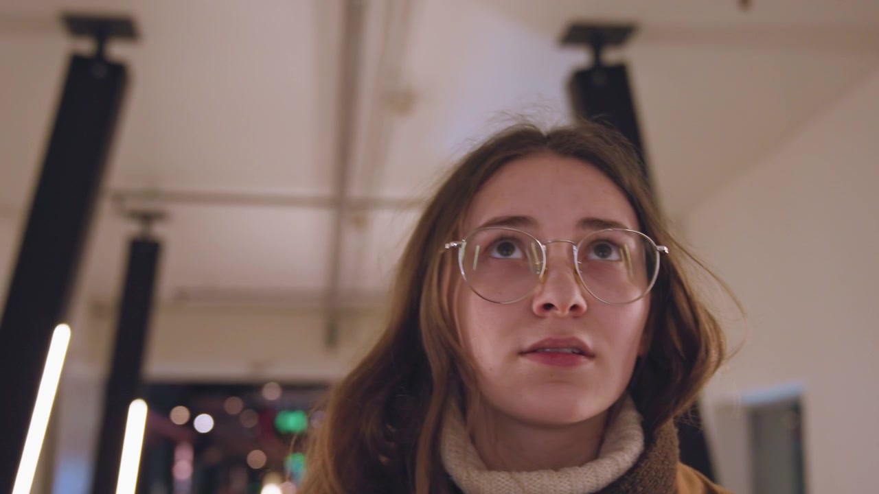 Close-up de mulher branca com expressão surpresa, jovem turista branca de óculos e casaco marrom reagindo a um visual repentino, sobrancelhas levantadas e boca ligeiramente aberta, luz de fundo LED suave, corredor interno.