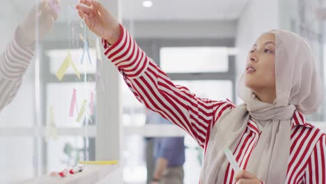 biracial businesswoman writing and sticking memo notes on glass blackboard at office, slow motion