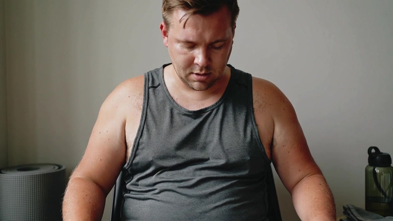 Hombre refrescándose y recuperando el aliento en una habitación pequeña con una botella de agua, vistiendo camiseta sin mangas gris y pantalones cortos negros.