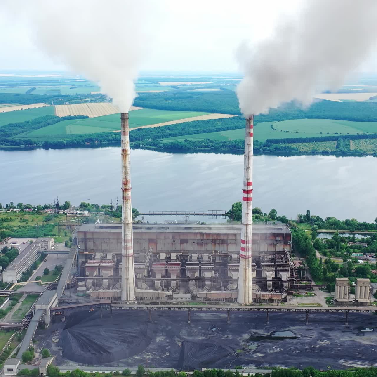 Factory with smokestacks near the river. High chimneys with thick emissions into the atmosphere on beautiful nature background. Air pollution. Aerial view