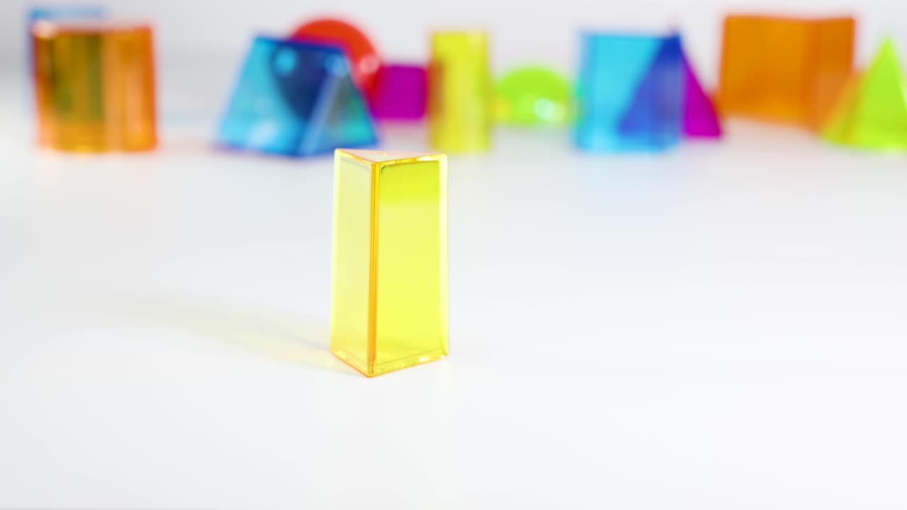 A hand arranges vibrant 3D geometric shapes on a white table, emphasizing color and form in a bright setting