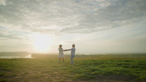 a couple joyfully spins on a grassy hill at sunset, next to a tranquil lake. the husband, in a white shirt and hat, embraces his wife in a flowing white dress