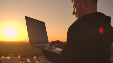 Close-up:-a-programmer's-hand-typing-on-a-laptop-keyboard-at-sunset-overlooking-the-roof.-A-businessman-works-remotely.-Freelancer-performs-work-on-vacation