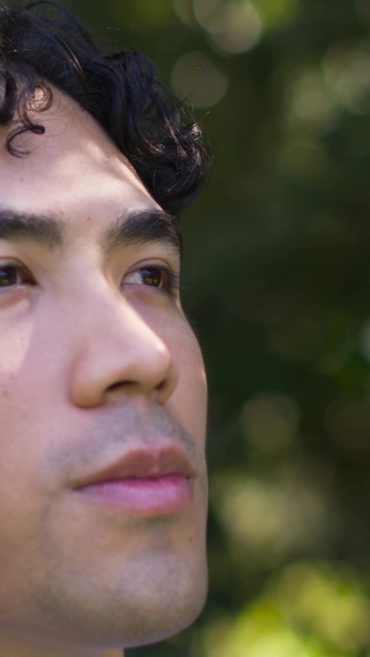 Video verticale: Uomo con capelli scuri che apre gli occhi al sole, alzando la testa in un parco frondoso, in camicia bianca.