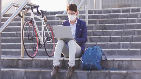 asian man wearing face mask using laptop while sitting on the stairs at corporate park