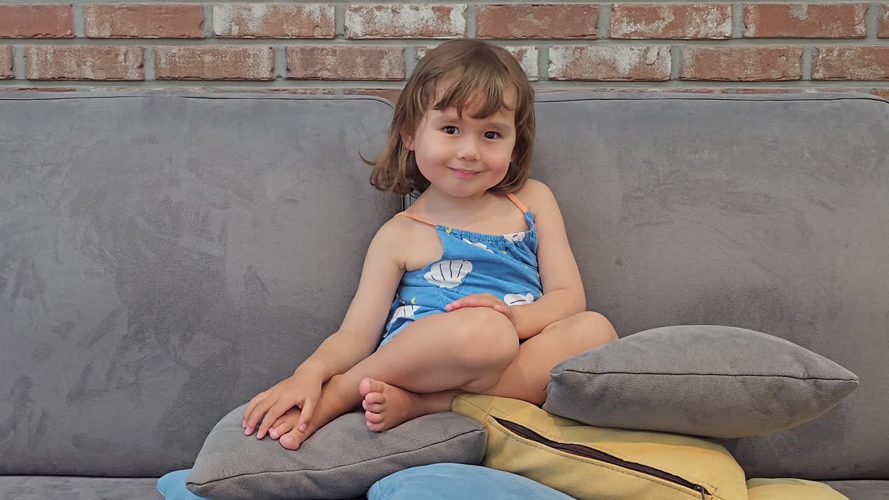 alegre niña de 3 años sentada en un sofá encima de almohadas de lanzamiento en casa