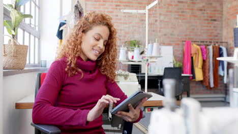 Happy-biracial-female-fashion-designer-sitting-and-using-tablet-in-studio,-slow-motion