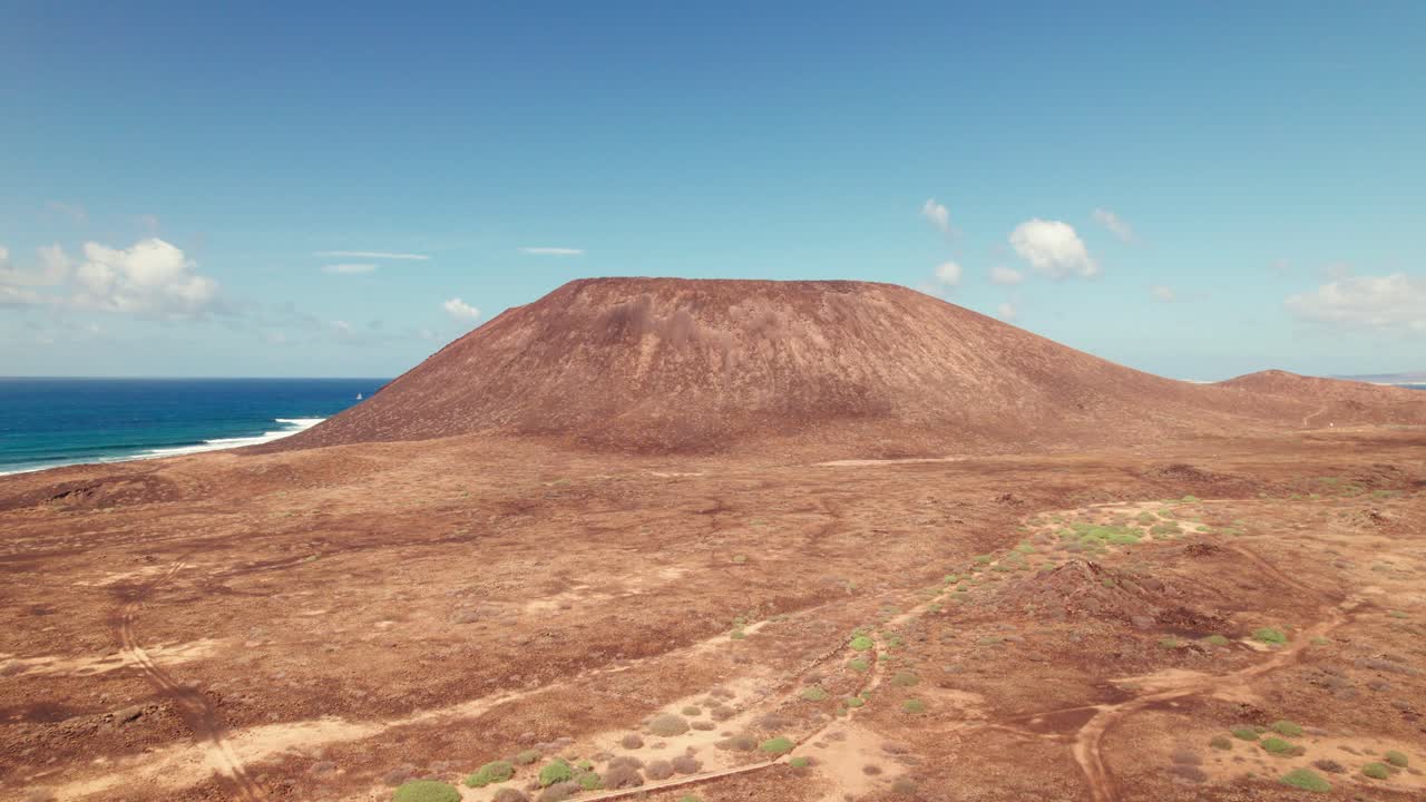 Aerial 4k drone view of Montaña de La Caldera on Isla de Lobos, Fuerteventura, showcasing the volcanic cone against a backdrop of ocean and clear blue sky.