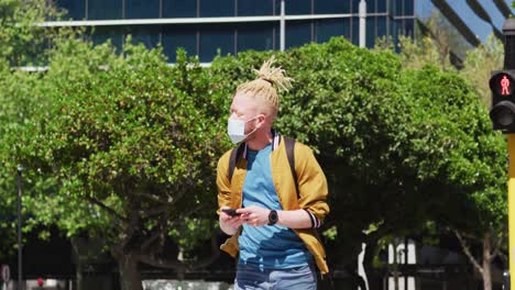 Albino-african-american-man-with-face-mask-and-dreadlocks-walking-and-using-smartphone