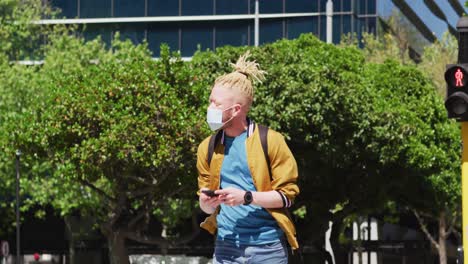 albino african american man with face mask and dreadlocks walking and using smartphone