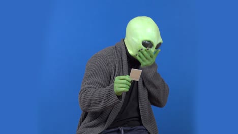 emotional alien with green skin in human clothes looks at picture and begins to cough sitting on blue chromakey background.