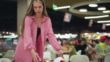 lady in pink dress carrying tray with burger, fries, and drink walks toward table, places it down, adjusts the tray, and sits with a focused expression, blurred background of people dining