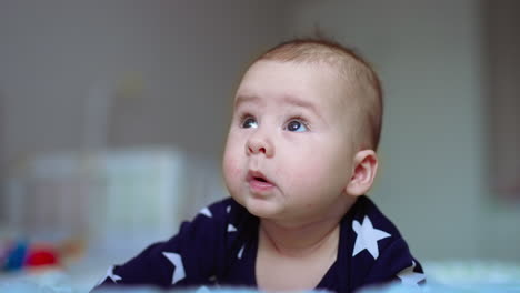 cute baby lies on bed on his stomach. beautiful infant child looks up with curiosity. close up. blurred backdrop.