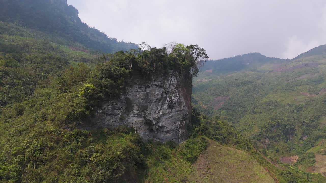 A majestic aerial view of a towering limestone cliff rising dramatically from a dense forest in Ha Giang, Vietnam. The sheer cliffs and lush foliage create a breathtaking contrast.
