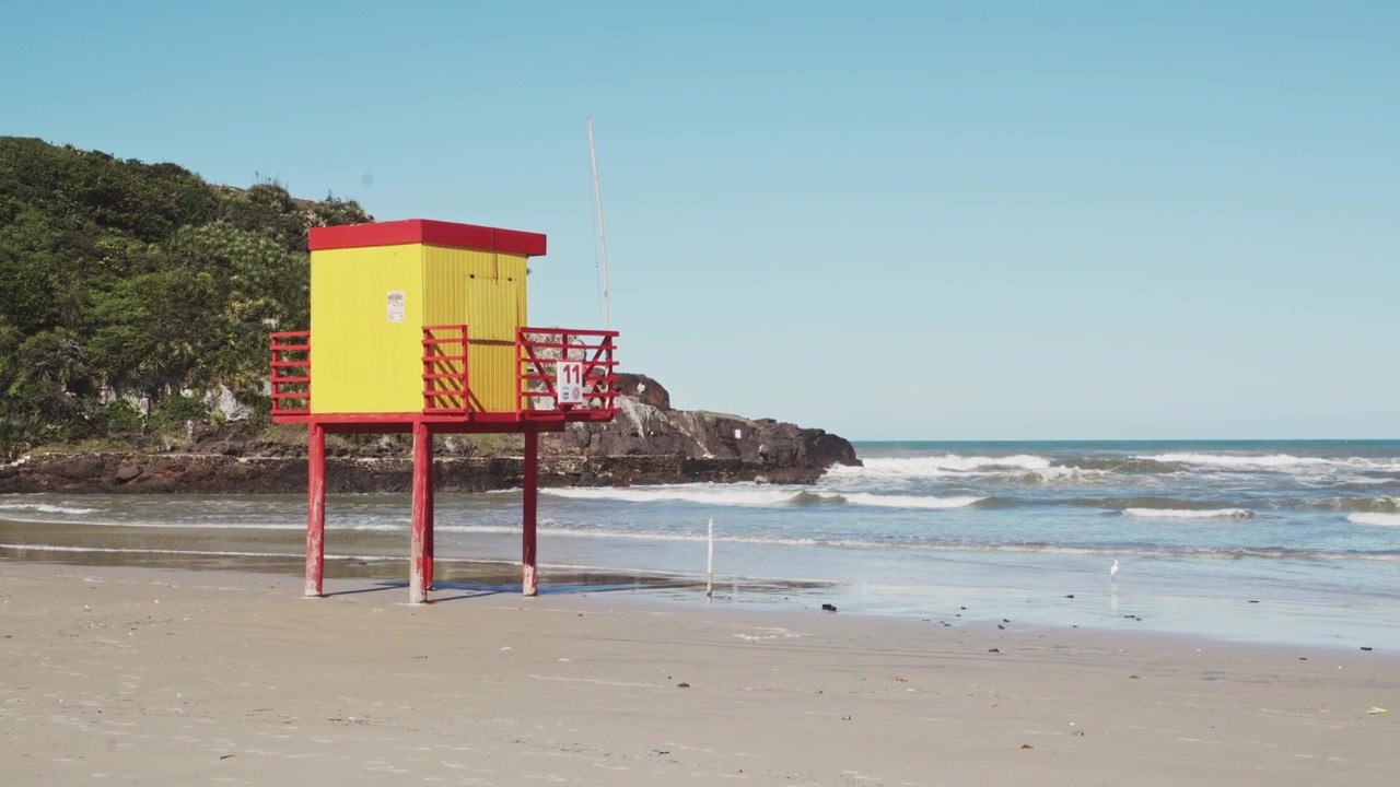 Un'inquadratura statica cattura una vivace torre di salvataggio gialla e rossa che si erge in modo prominente su una spiaggia sabbiosa. Le onde si infrangono dolcemente sullo sfondo, creando una serena scena costiera perfetta per un viaggio sicuro.
