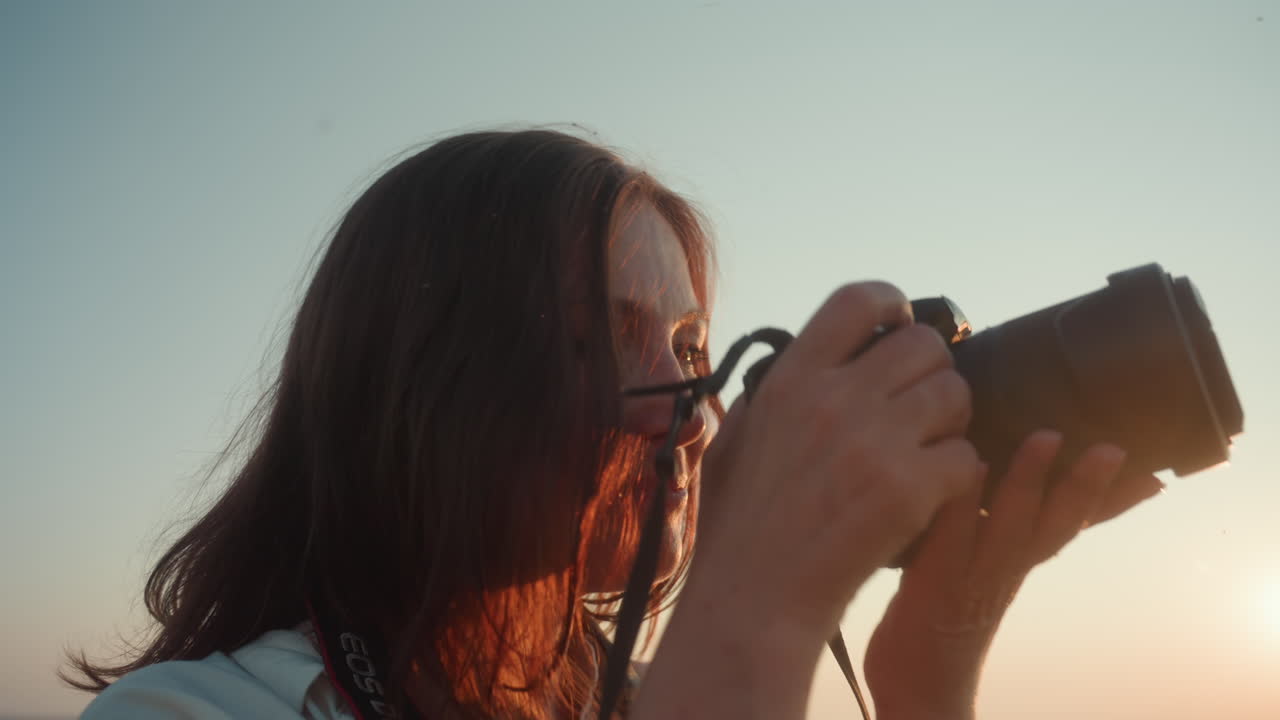 Close up of woman holding camera to eye as she takes photo during golden hour, soft sunlight glowing on her hair and face, capturing peaceful moment with calm expression