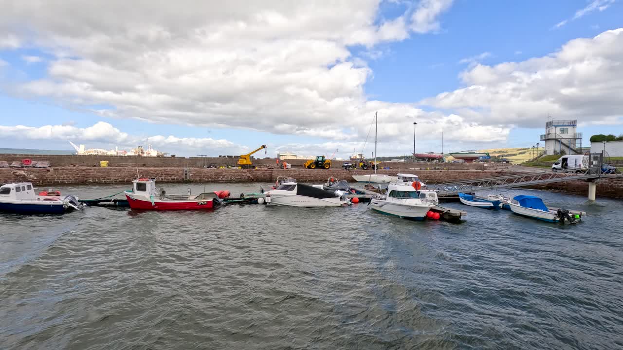 Small fishing boats and motorboats sway on choppy water beside a pier in Portree harbor, under bright daylight with partly cloudy skies. Subtle camera movement captures the dynamic coastal atmosphere