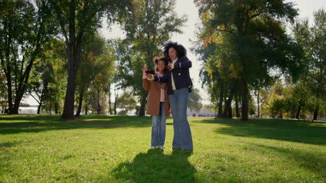 Madre-Feliz-Haciendo-Llamadas-En-Línea-Con-Una-Linda-Hija-Sonriente-Parada-En-El-Parque.