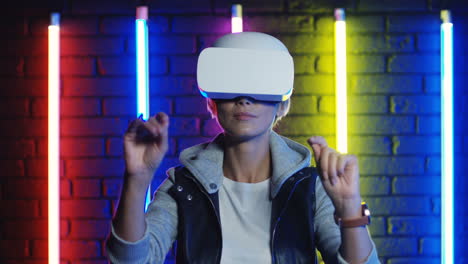 close up view of young blonde woman in vr glasses moving her hands in the air in a room with colorful neon lamps on the wall