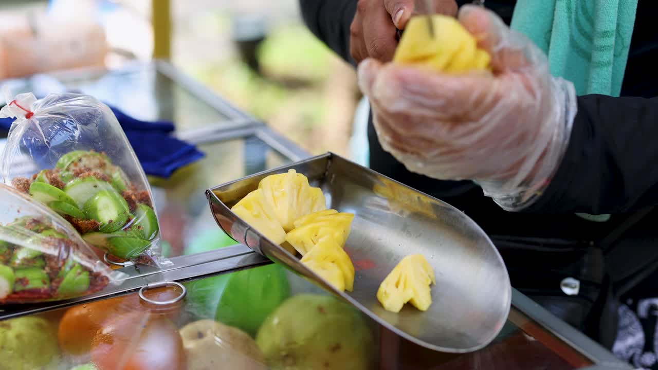 Gloved vendor slices fresh pineapple at outdoor fruit cart, natural daylight, close-up handheld shot