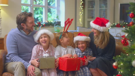 Family-Wearing-Santa-Hats-Sitting-On-Sofa-At-Home-Opening-Christmas-Gifts