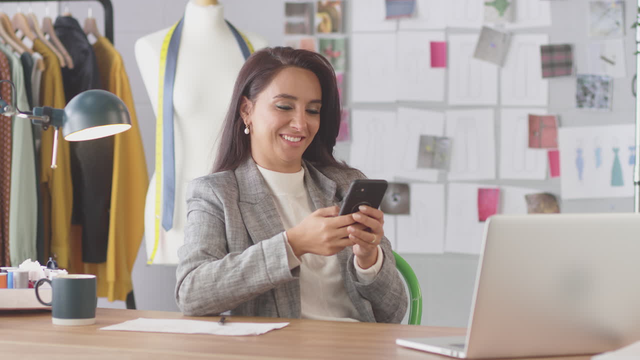 diseñadora de moda en el estudio trabajando en el escritorio en la computadora portátil y comprobando mensajes en el teléfono móvil