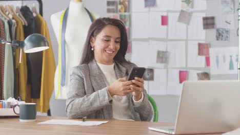 diseñadora de moda en el estudio trabajando en el escritorio en la computadora portátil y comprobando mensajes en el teléfono móvil