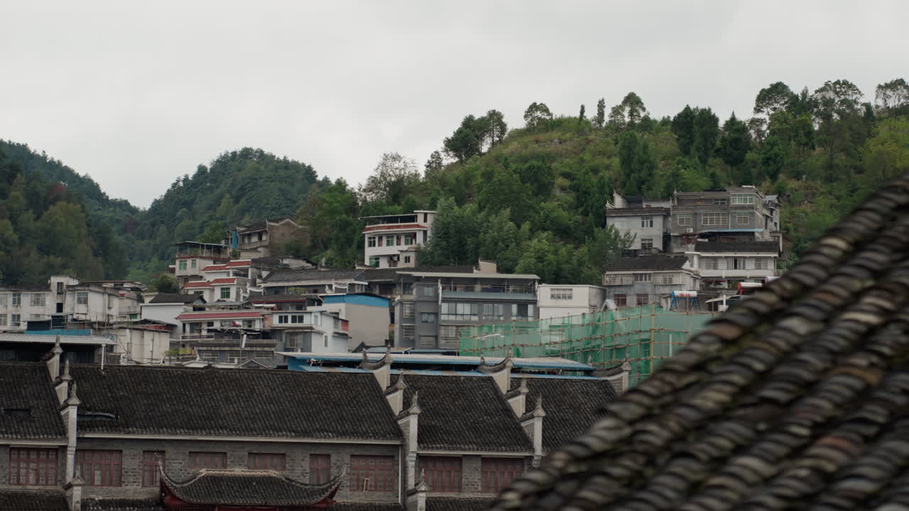 Ngôi làng trên sườn đồi u ám với mái ngói, tầm nhìn toàn cảnh các tòa nhà pha trộn truyền thống và hiện đại nép mình bên những ngọn núi xanh, có thể nhìn thấy giàn giáo và kết cấu mái nhà dày đặc, màu sắc trầm.