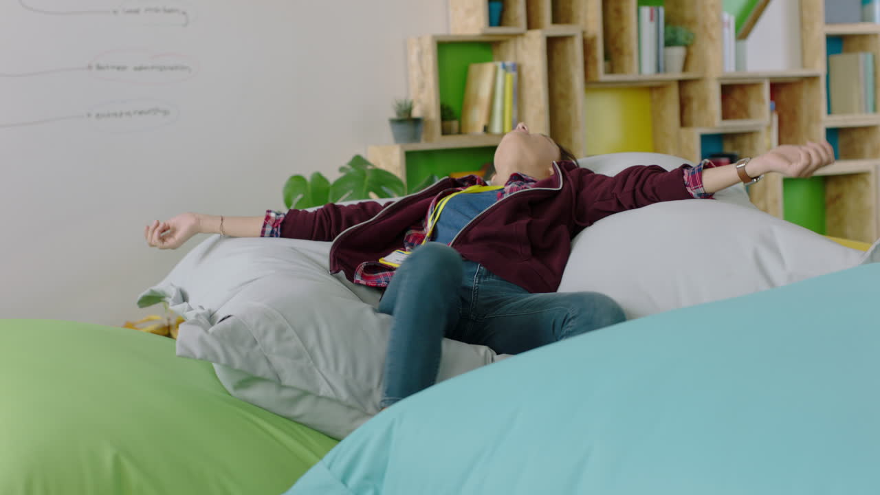 행복한 백인 여성 학생이 다채로운 베개에 뛰어들고 여자가 휴식을 취하고 트렌디한 사무실 작업장에서 점심 휴식을 즐기고 있습니다.