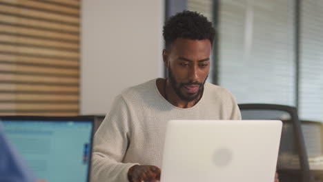 two businessmen working at desks on laptops discussing document on screen in modern office