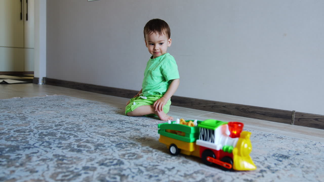 Beautiful dark-haired toddler sits on the floor. Lovely child watches how the toy car moves by the carpet.