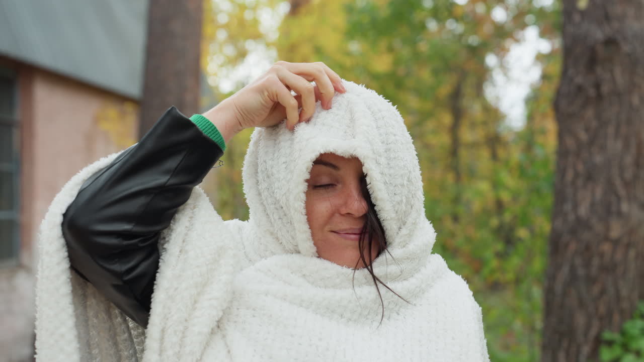 Mulher branca com capa de tricô puxa suavemente o capuz para uma revelação divertida, manga de couro à mostra, cabana rústica e pinheiros emoldurando uma cena de outono, tricô texturizado, sorriso espontâneo, retrato aconchegante ao ar livre