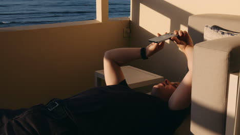 boy lying down holding phone in hands, hotel balcony in sunlight with sea view, hawaii