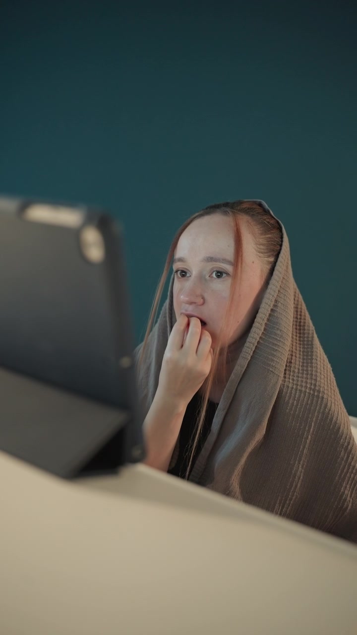 Woman sits at table wrapped in blanket and eating food. Woman holds object near her mouth with minimal movement throughout. Scene is indoors against teal wall background
