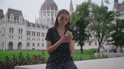 young woman using phone in front of the hungarian parliament building