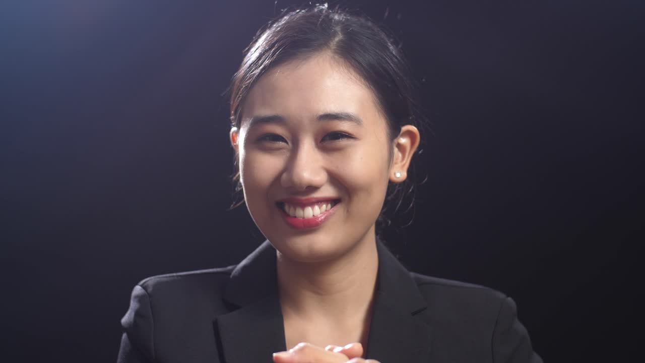 블랙 스크린 스튜디오에 서서 손을 박고 주변을 둘러보는 미소 짓는 아시아 스피커 여성