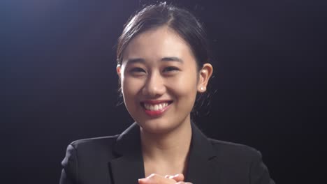 close up of smiling asian speaker woman in business suit clapping her hands and looking around while standing in the black screen studio